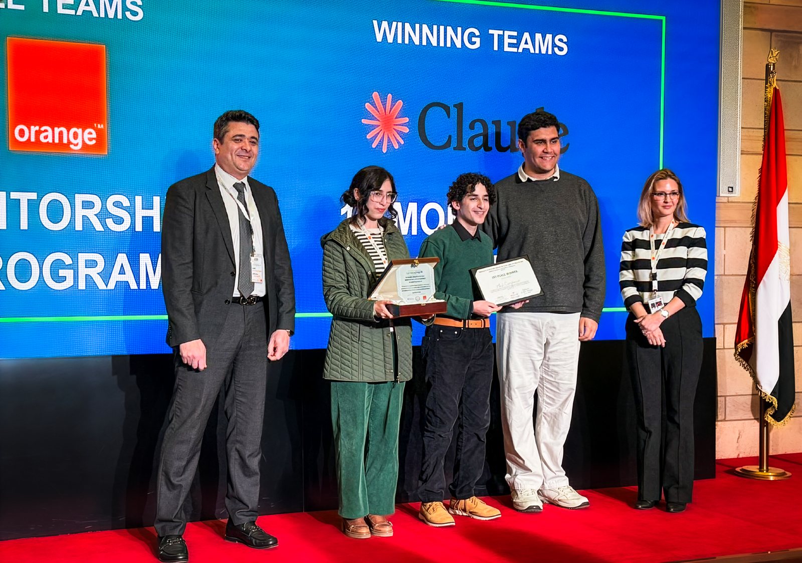 Gamal Sheridan on stage with two MediCompanion teammates, holding the first place certificate and plaque at the Winning Teams ceremony, beneath a screen showing the Orange and Claude logos